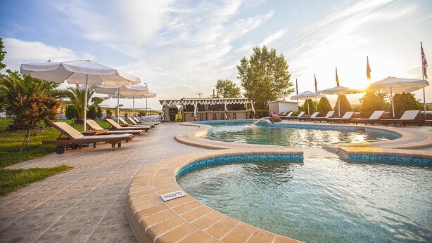 Ein Swimmingpool mit Liegestühlen und Sonnenschirmen, umgeben von einer gefliesten Terrasse und Grünflächen.