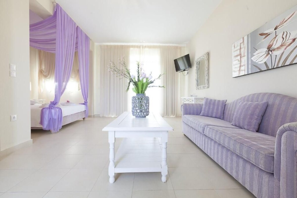 A room with a bed, a white coffee table, a purple sofa, a TV, and a painting.