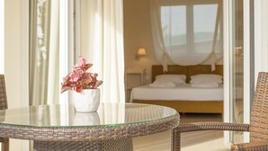 Ein Hotelzimmer mit einem Bett, einem Glastisch mit einer Topfpflanze, Korbstühlen und Blick auf das Meer durch das Fenster.