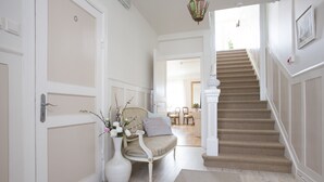 En gang med beige tæppe, en hvid dør, en hvid vase med blomster, en stribet sofa og en trappe med beige tæppe, der fører til et andet rum.