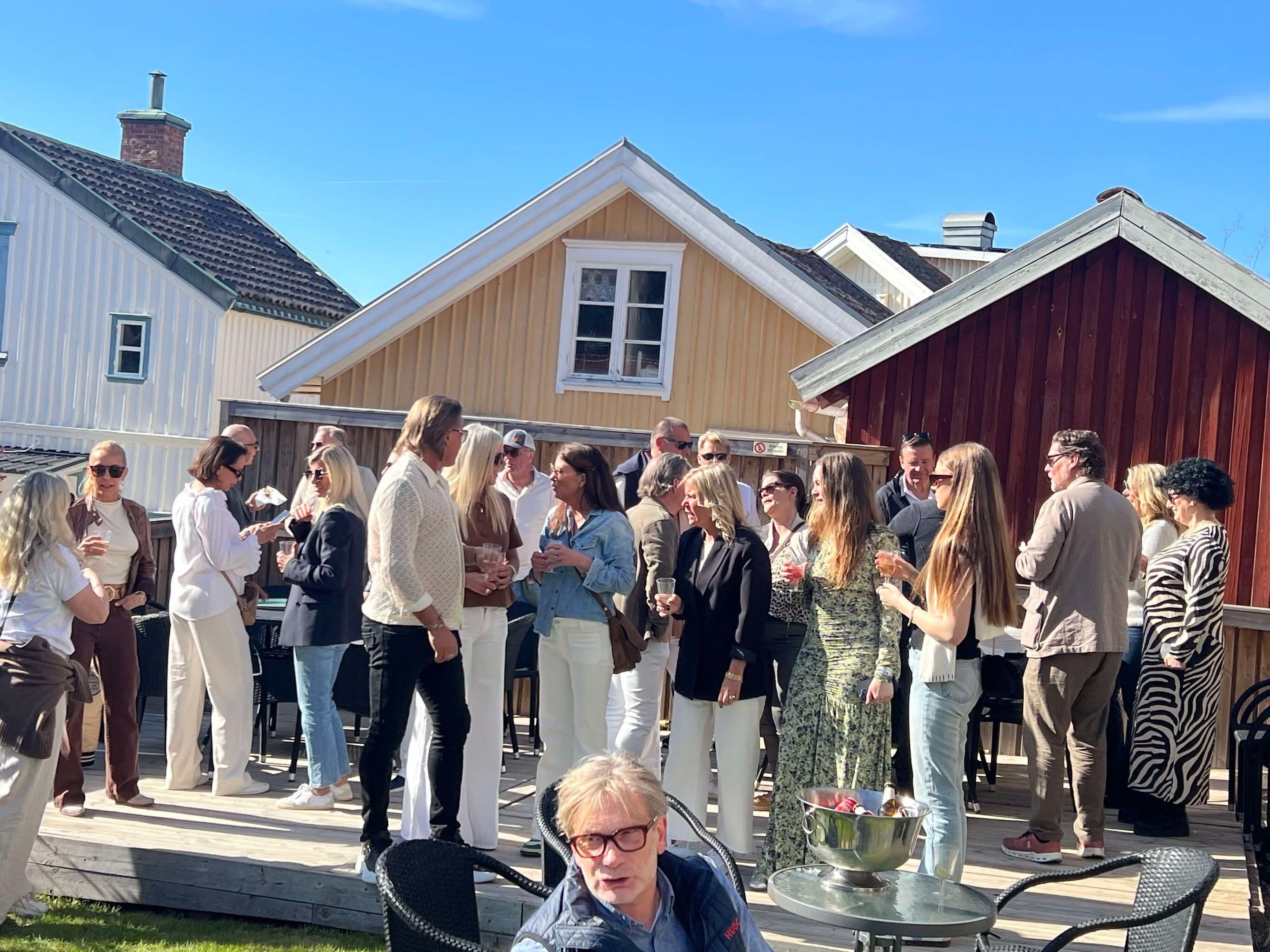 A group of people gathered outdoors, possibly at a social event, with a wooden building and a red barn in the background.