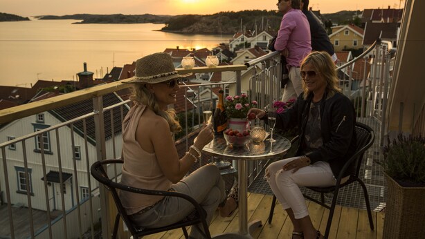 En balkon med udsigt til solnedgangen over vandet, et bord med drikkevarer og en kurvestol.'