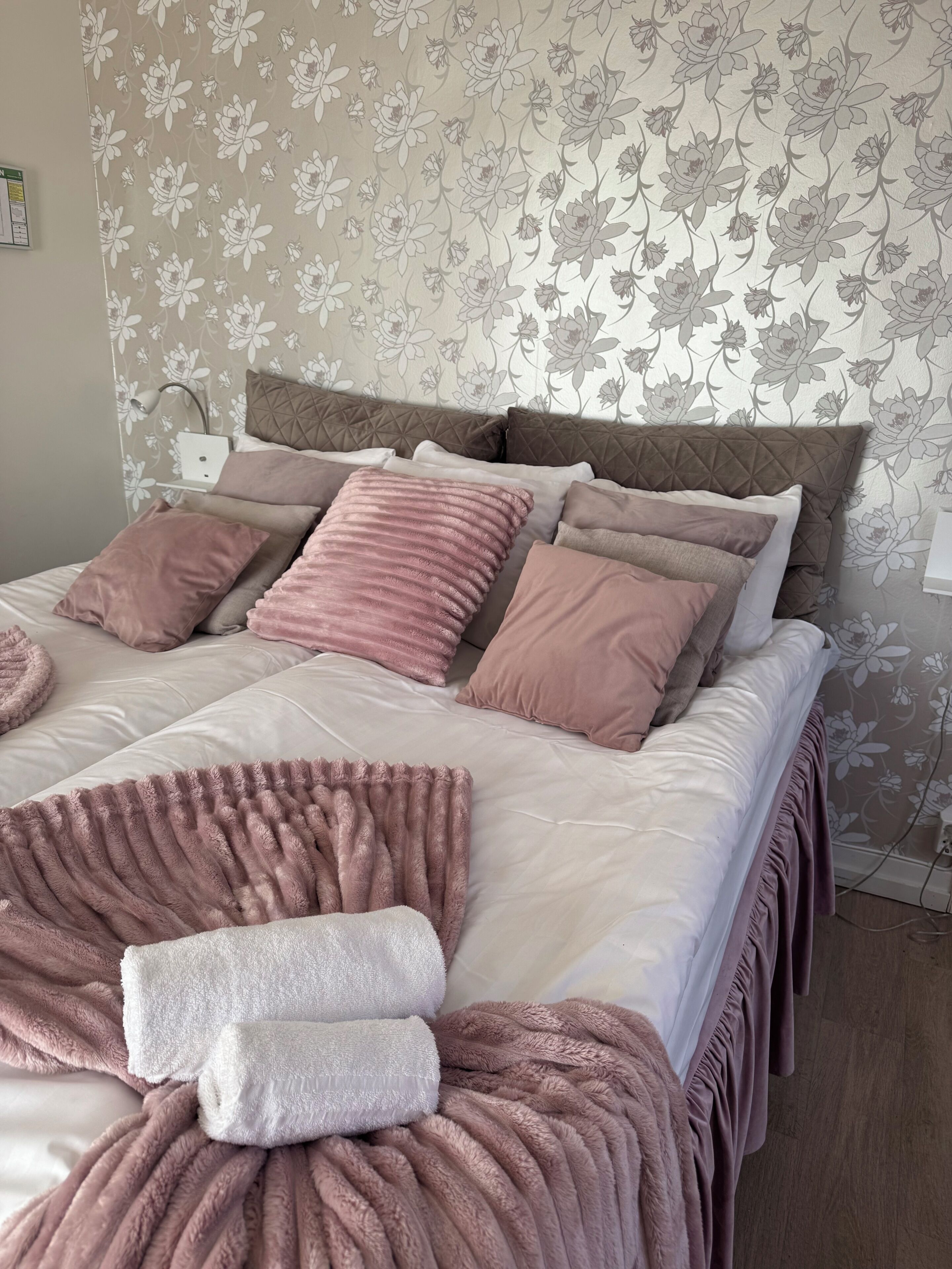 A neatly made bed with multiple pillows and a blanket, a bedside lamp, and a floral wallpaper pattern on the wall.