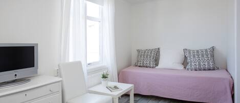 Une chambre à coucher avec un lit, une télévision, une chaise, une petite table et une fenêtre avec des rideaux.