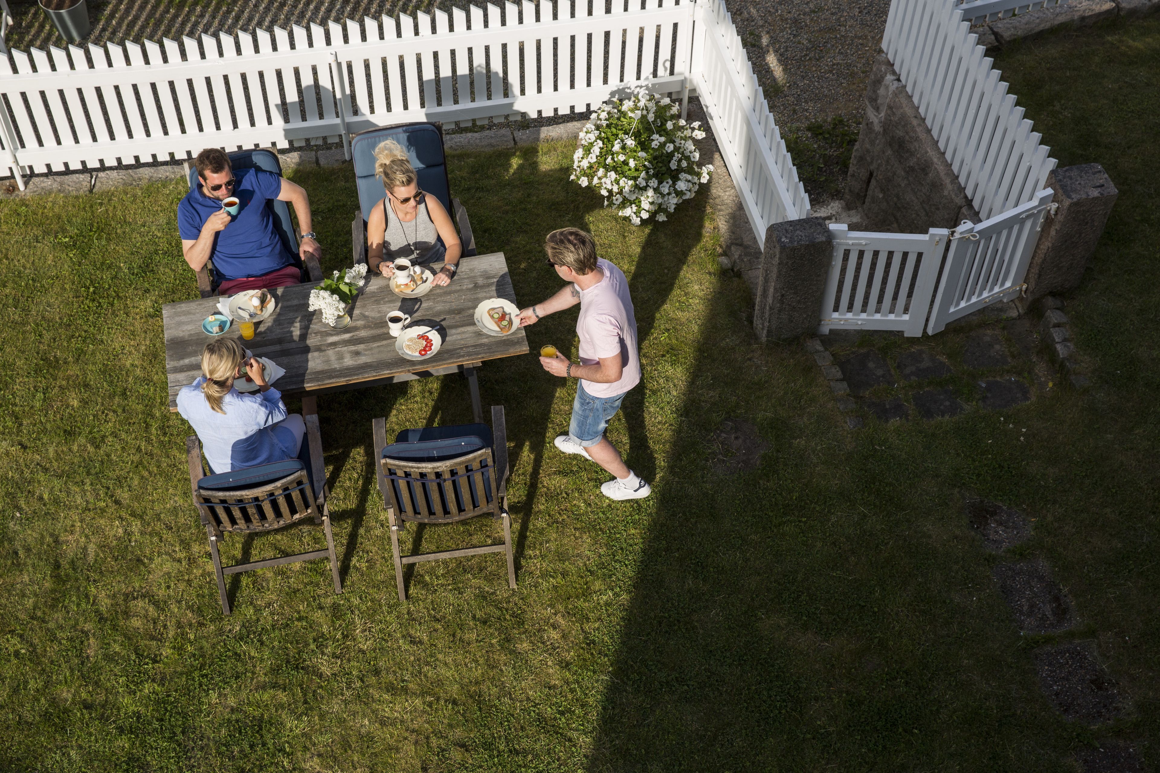 A group of people dining outdoors at a wooden table with chairs, surrounded by a white picket fence and a garden area.