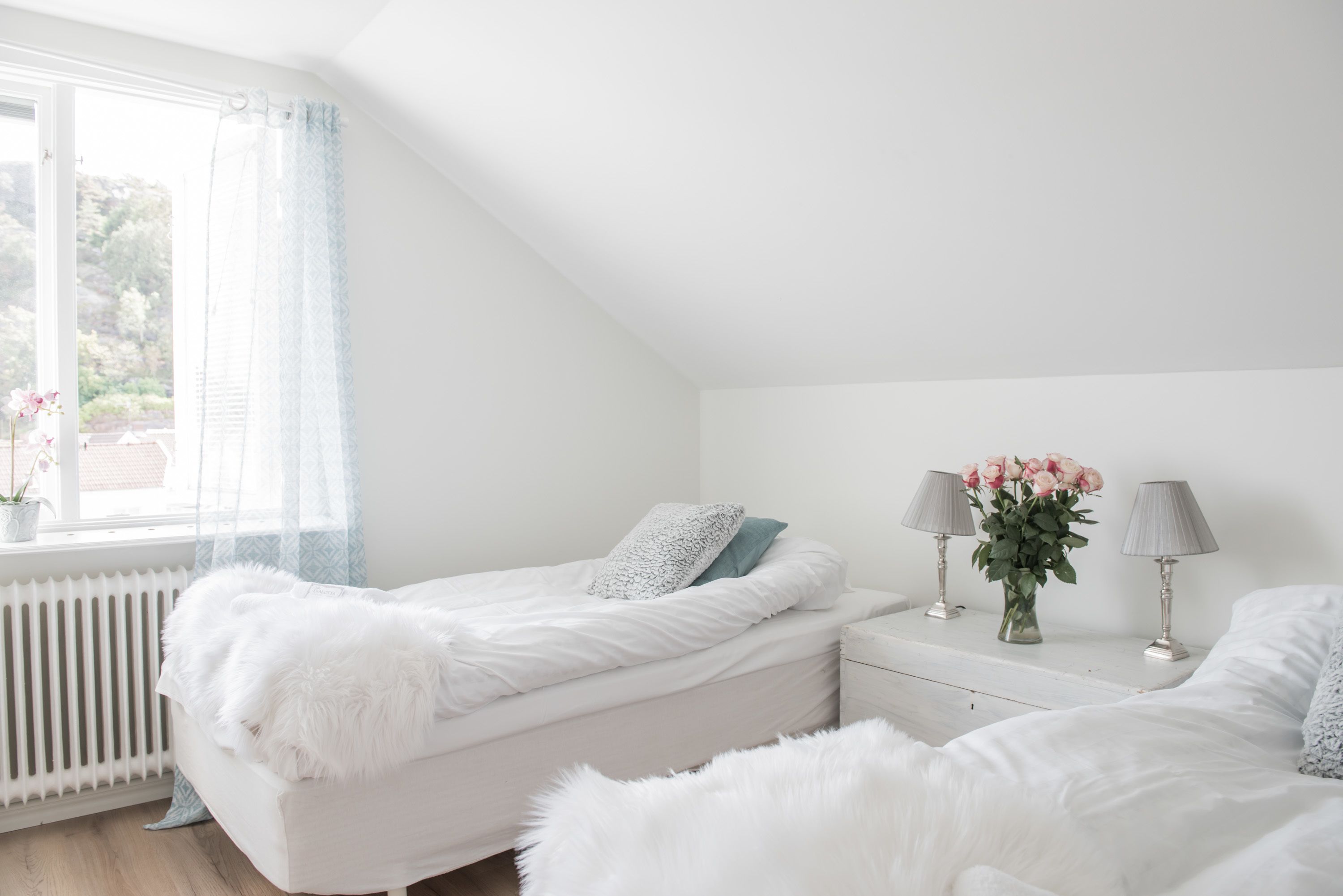 A bedroom with two beds, a window with curtains, a bedside table with lamps, and a radiator.