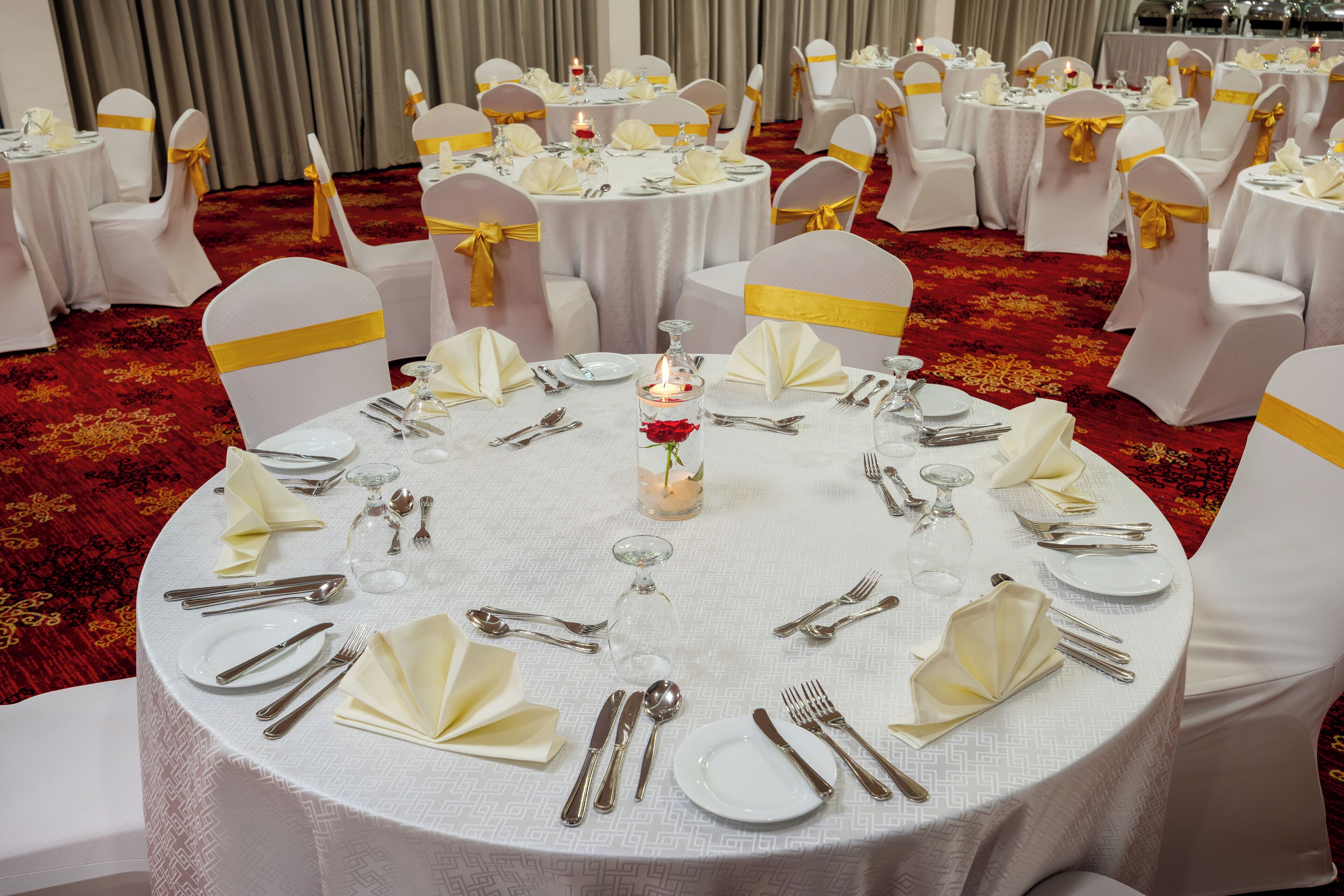 A banquet hall with round tables set for a formal event, each with white tablecloths, silverware, and yellow sashes on chairs.