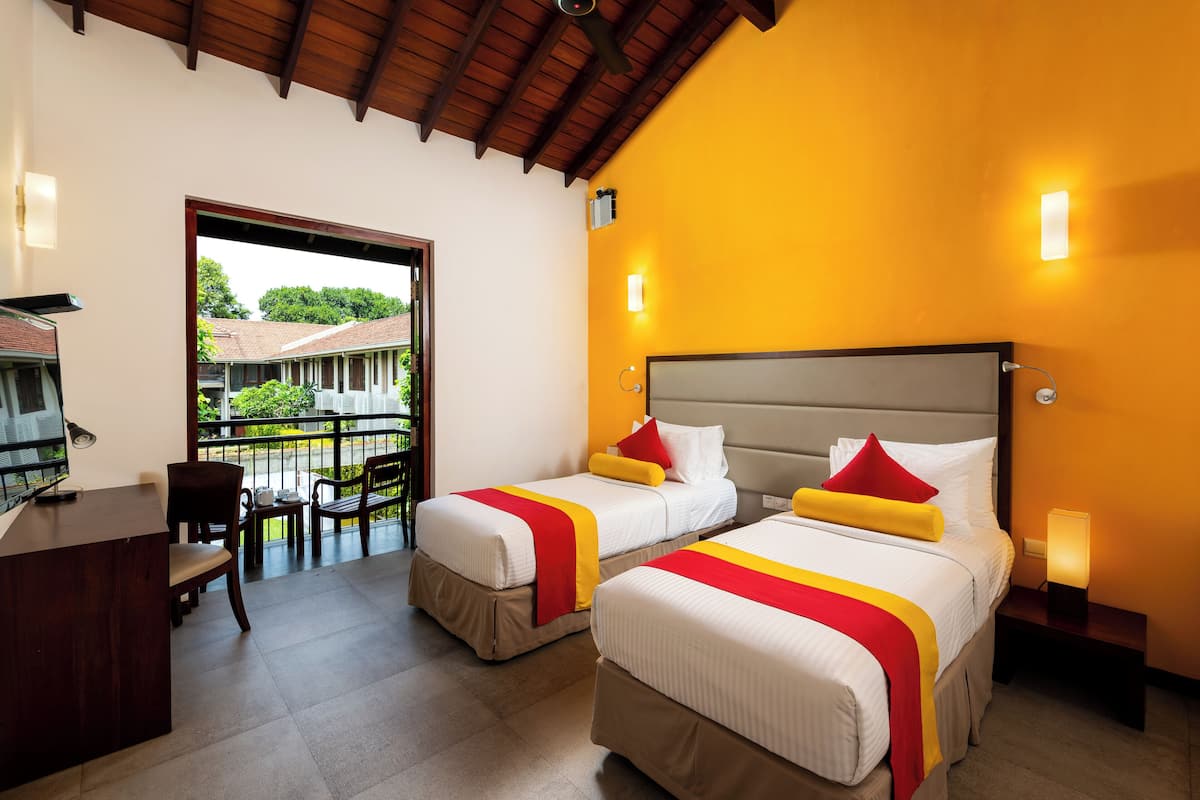 A hotel room with two beds, a desk, and a balcony with a view of greenery.