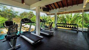 A gym with treadmills, a stationary bike, and dumbbells, with a view of lush greenery and a pool outside.