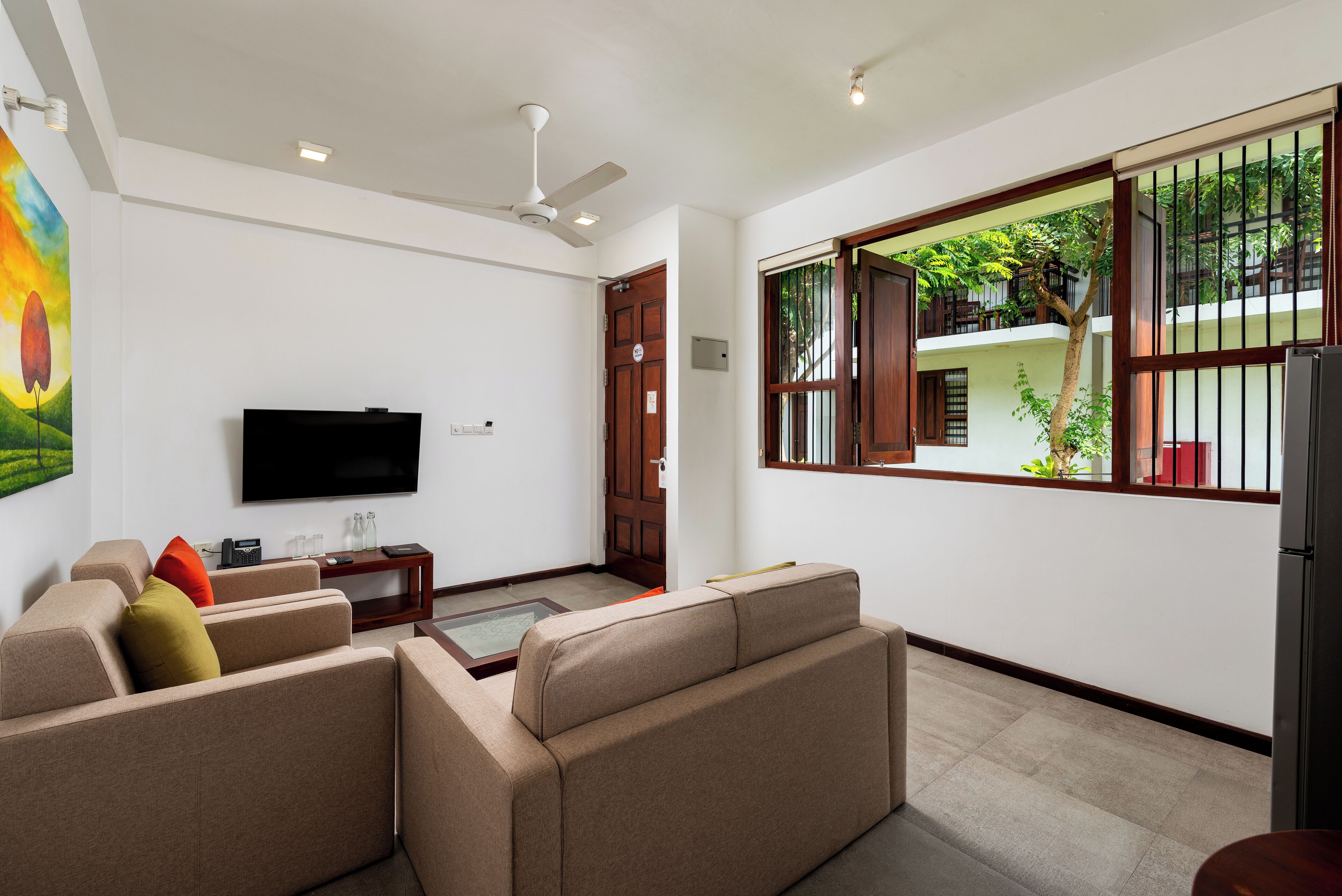 A modern living room with a flat-screen TV, beige sofas, a coffee table, and a large window with a view of a balcony.