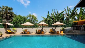 A swimming pool with sun loungers and umbrellas, surrounded by lush greenery and a clear blue sky.