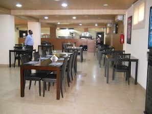 Un grand espace dédié à la salle à manger, avec plusieurs tables et chaises, une cuisine à l’arrière-plan et un extincteur fixé au mur.