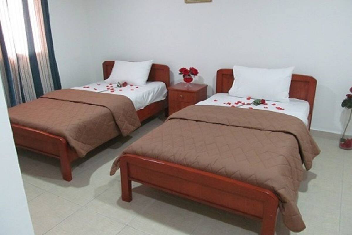 Une chambre d’hôtel avec deux lits simples, chacun doté d’un cadre en bois et d’une literie marron. Une petite table de chevet en bois, sur laquelle est posé un vase de fleurs rouges, se trouve entre les deux lits.