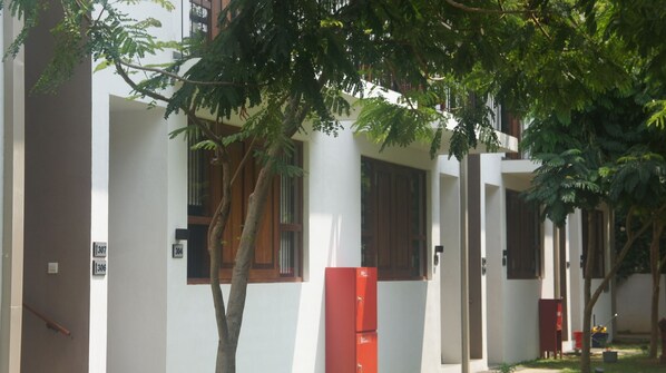 Une rangée de bâtiments blancs avec des portes numérotées, des arbres au premier plan et un objet rouge sur le sol.