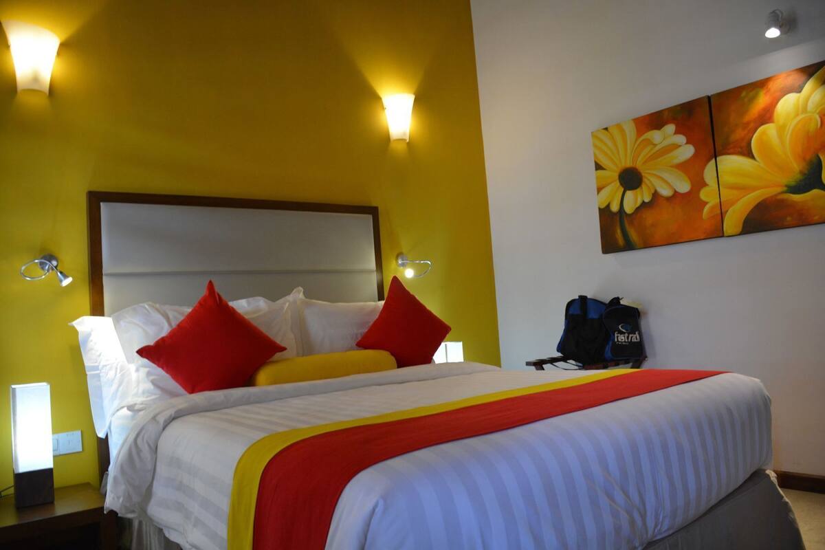 A hotel room with a bed, red and white pillows, a yellow wall, and a painting of flowers.