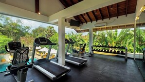 A gym with treadmills, an elliptical, and dumbbells, with a view of lush greenery and a pool outside. - The Thinnai Hotel (Jaffna)