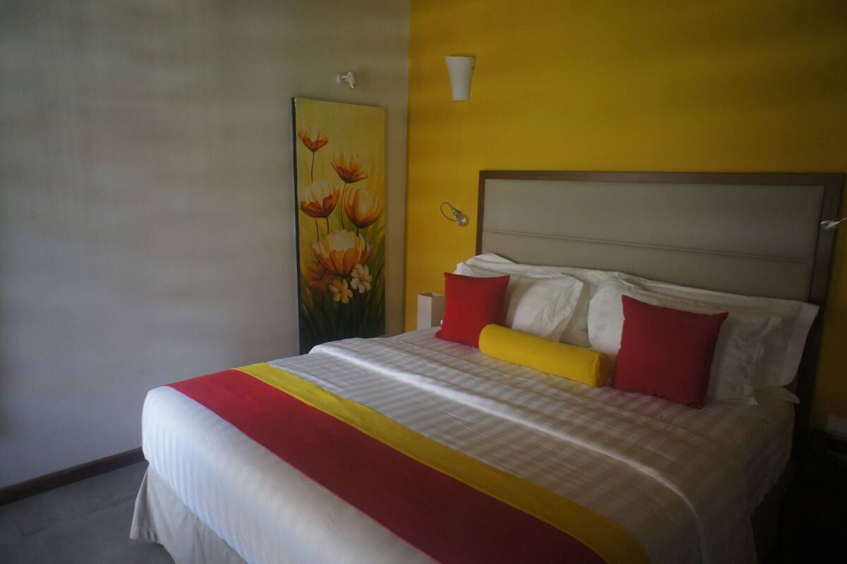 A bed with a white headboard, red and yellow pillows, and a painting of flowers on the wall.