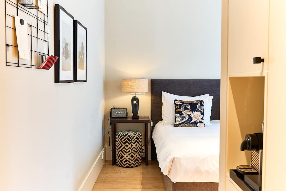 A hotel room with a bed, a nightstand with a lamp, a wall-mounted shelf with papers, and a patterned ottoman.