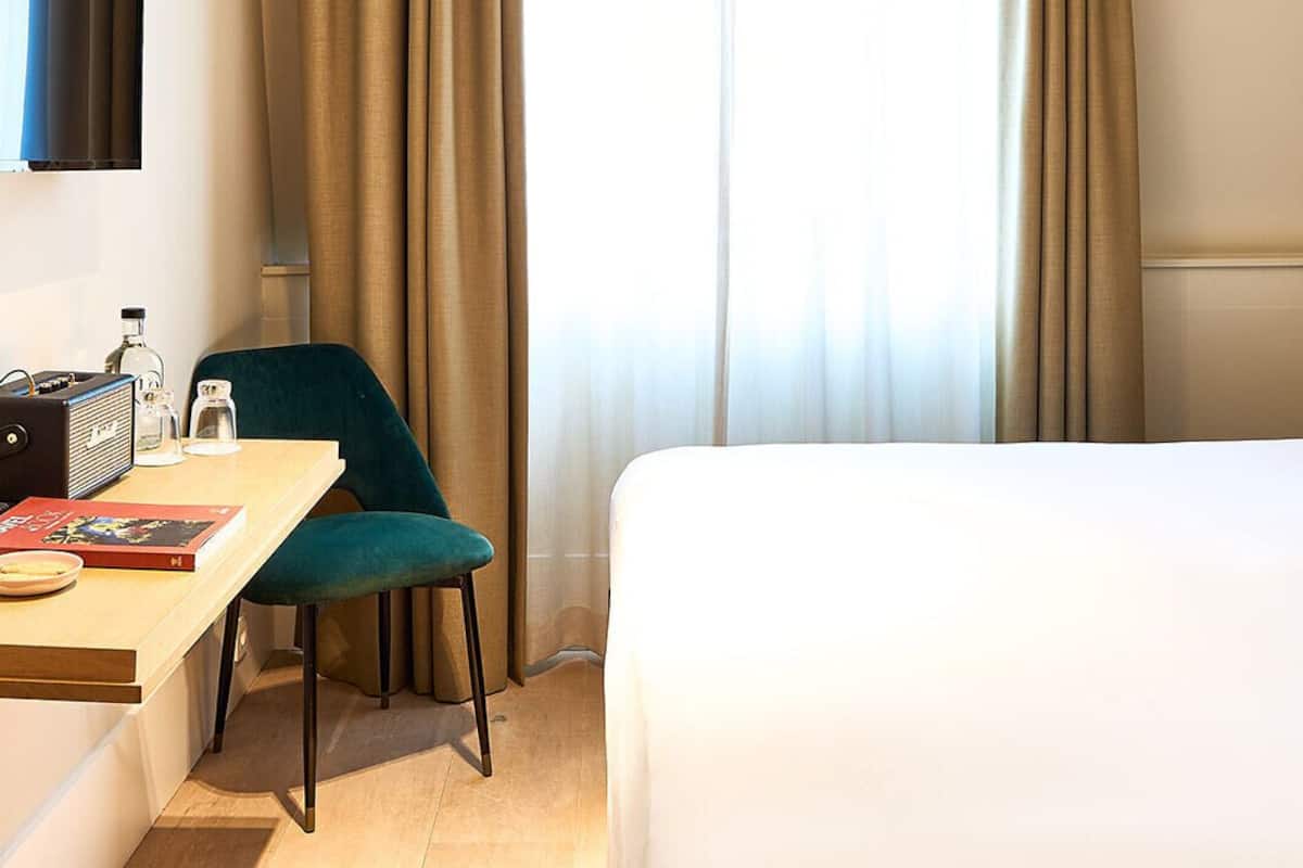 A hotel room with a bed, a desk with a chair, a flat-screen TV, and a window with curtains.