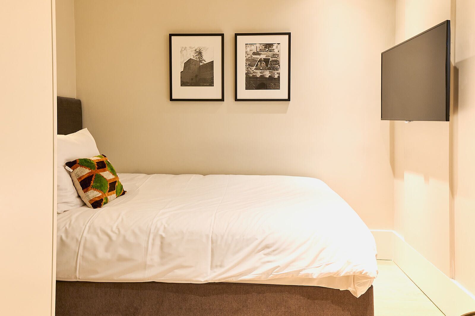 Un lit bien fait, recouvert d’une couette blanche et agrémenté d’un coussin aux motifs géométriques colorés, deux photographies encadrées accrochées au mur et un téléviseur à écran plat fixé au mur.