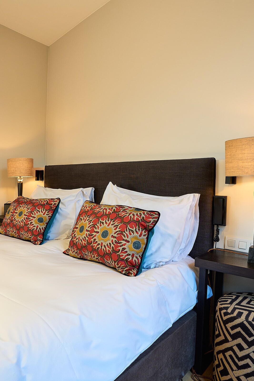 Una cama bien hecha con sábanas blancas y dos almohadones decorativos, un cabecero oscuro, lámparas de noche y una mesita con un teléfono y una lámpara.