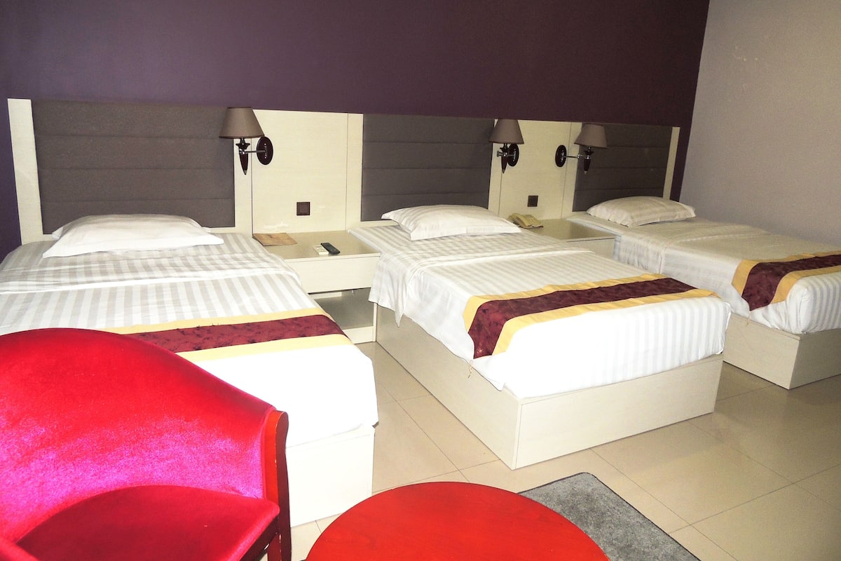 A hotel room with four beds, each with a bedside table and lamp. There is a red chair and a small round table in the foreground.