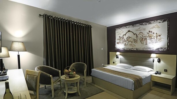 Une chambre d’hôtel avec un lit, deux chaises, une petite table sur laquelle se trouve un panier de fruits, une lampe et un tableau au mur.