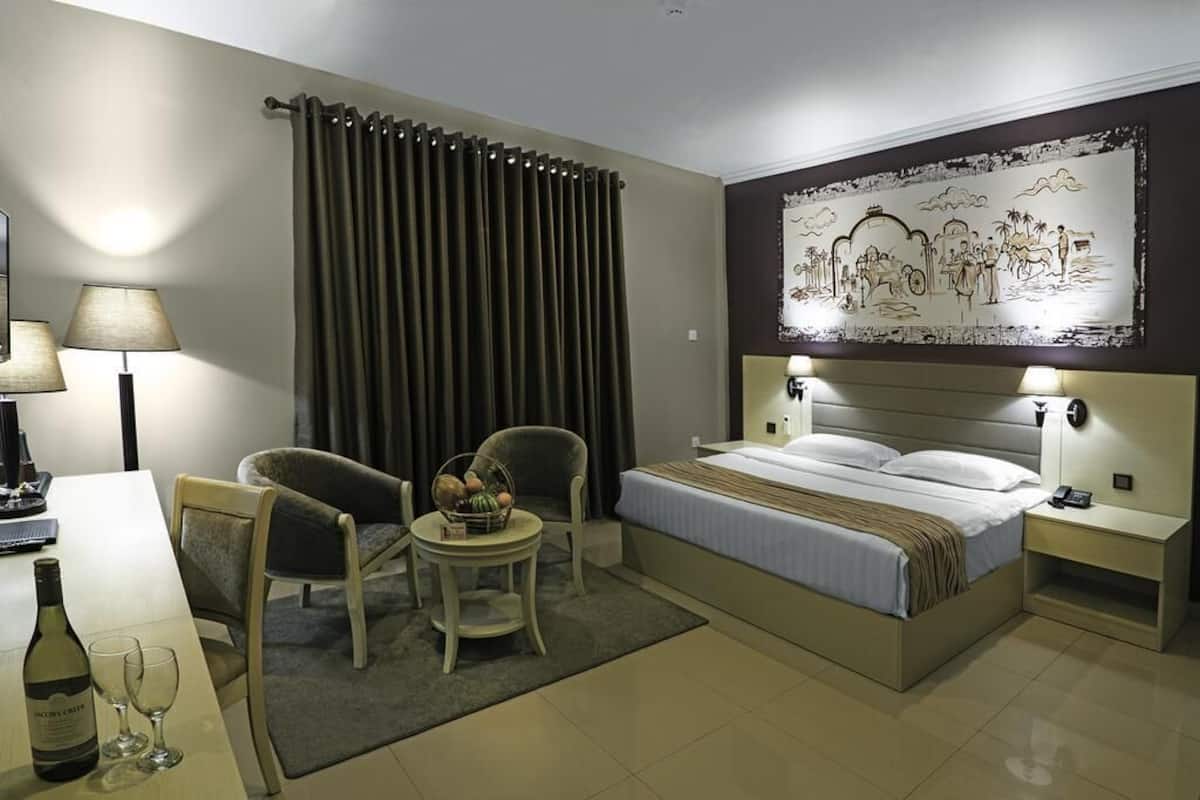 A hotel room with a bed, two chairs, a small table with a fruit basket, a lamp, and a painting on the wall.