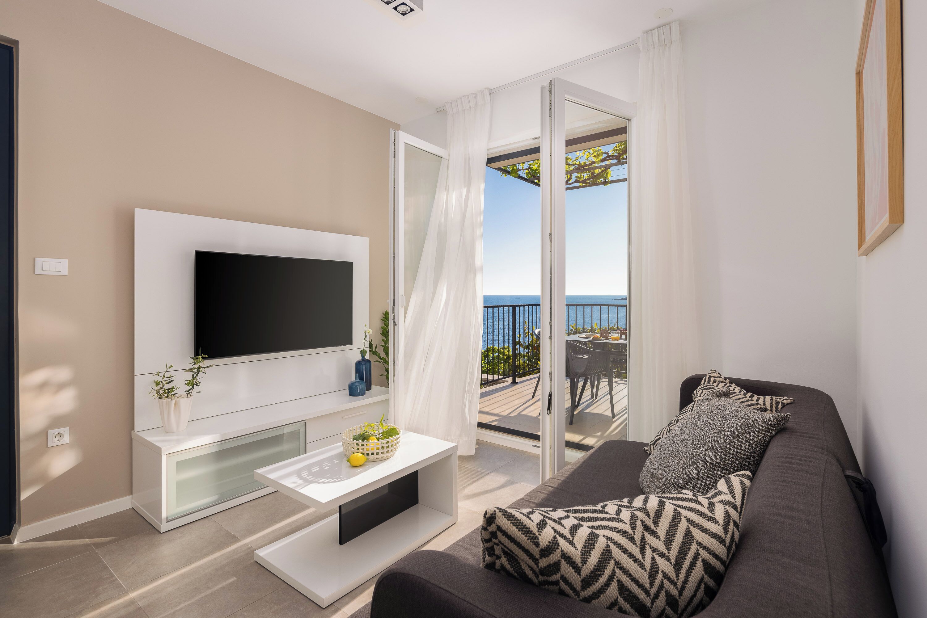 Ein modernes Wohnzimmer mit einem Flachbildfernseher, einer weißen Konsole, einem grauen Sofa mit gemusterten Kissen und einem Balkon mit Meerblick.