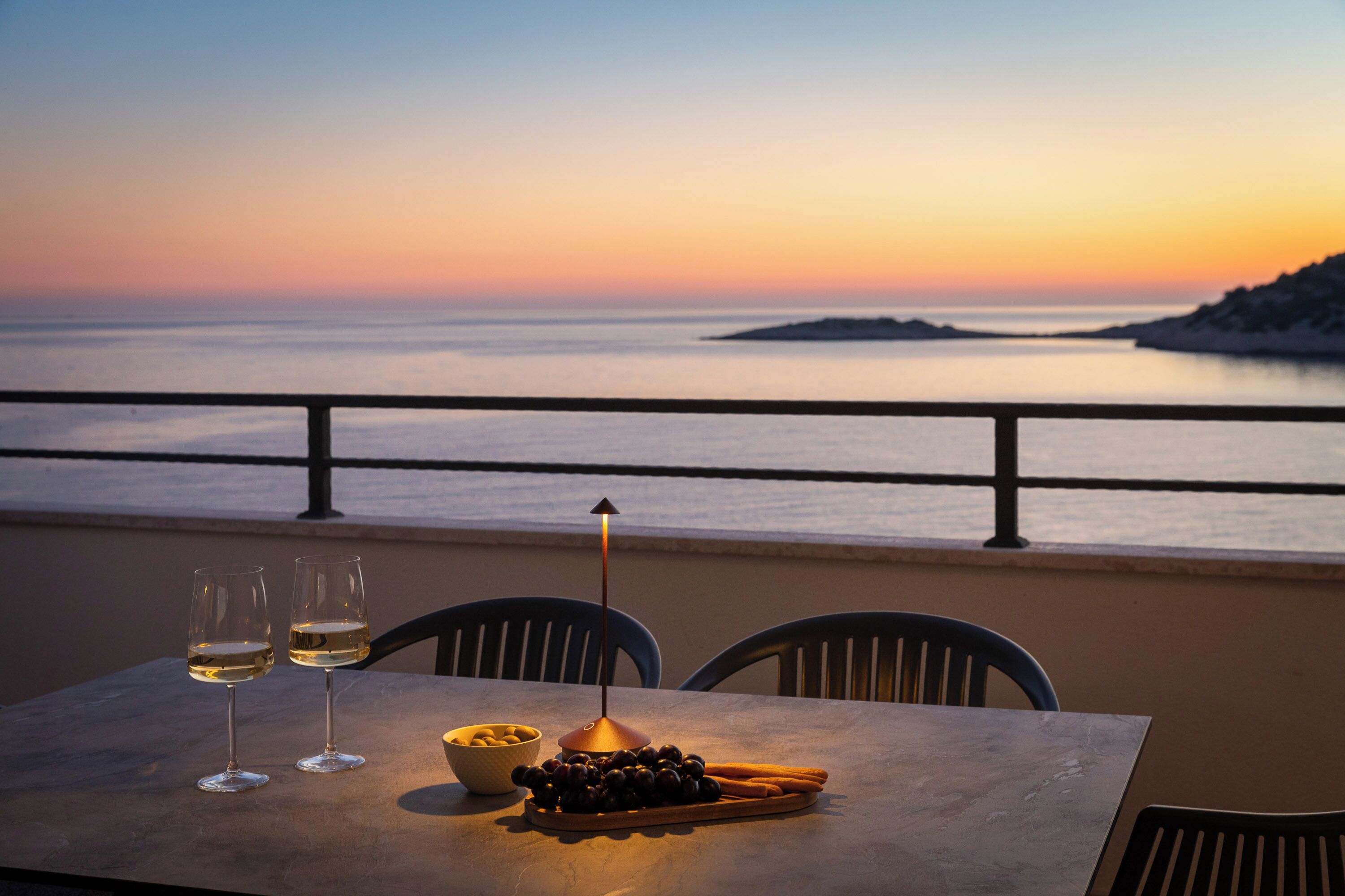 Una mesa con dos copas de vino blanco, un cuenco de aceitunas y una bandeja de palitos de pan en un balcón con vista a la puesta de sol sobre el mar.
