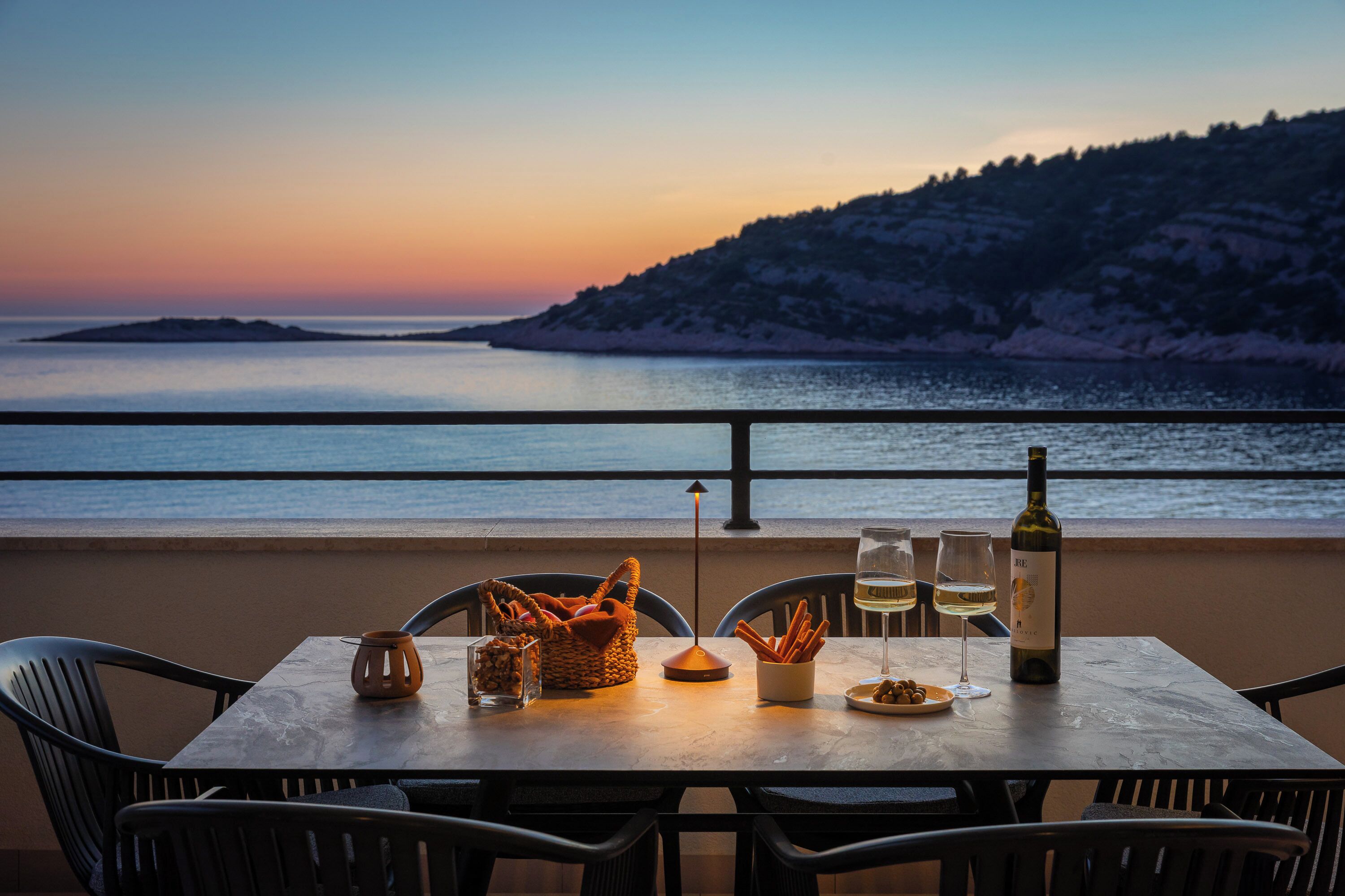 Un tavolo apparecchiato con una bottiglia di vino, bicchieri, un cesto e un piatto di stuzzichini su una terrazza con vista su un corpo d'acqua e un paesaggio collinare.