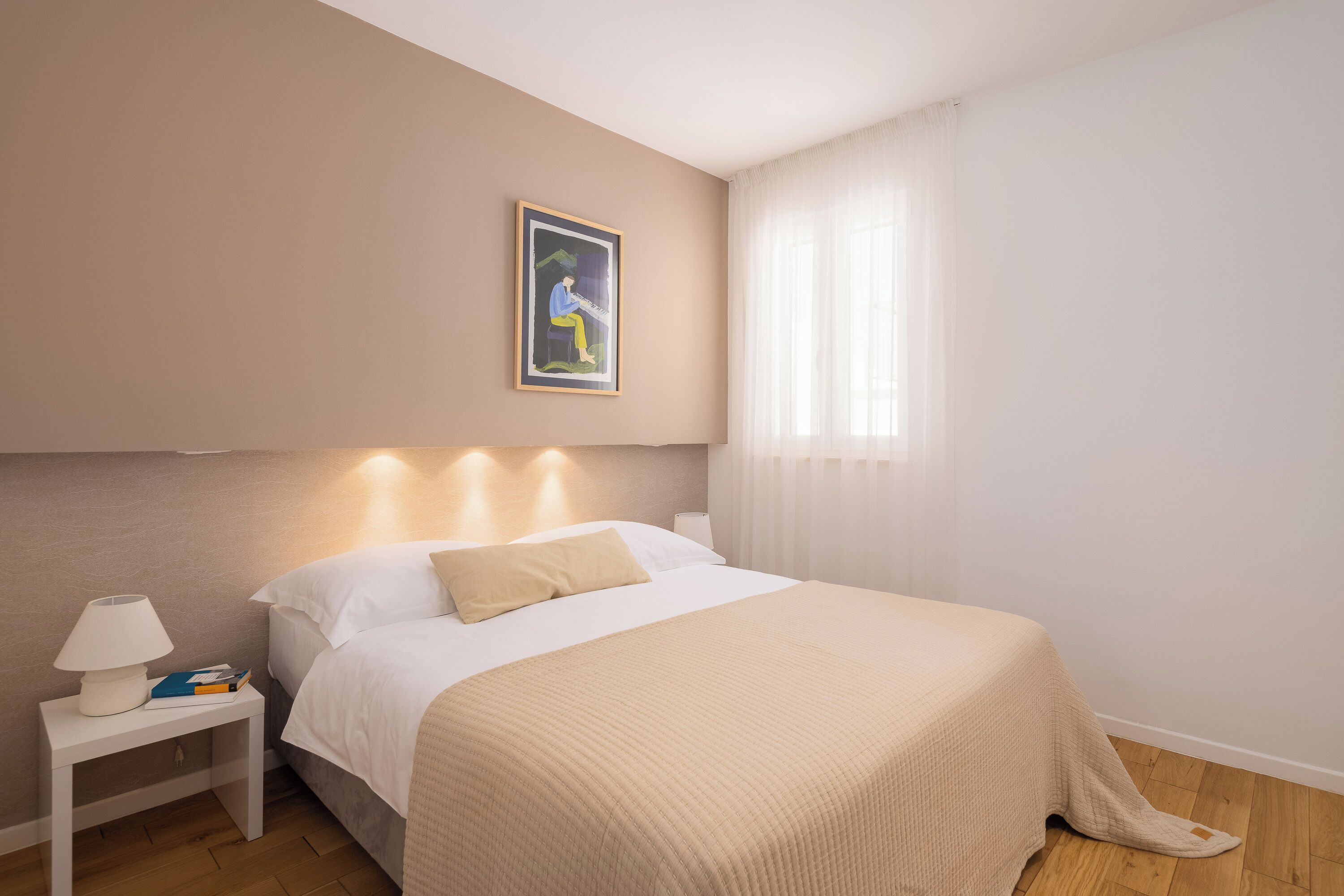 Une chambre d’hôtel bien rangée, avec un lit, une table de chevet surmontée d’une lampe, un tableau encadré au mur et des rideaux légers à la fenêtre.