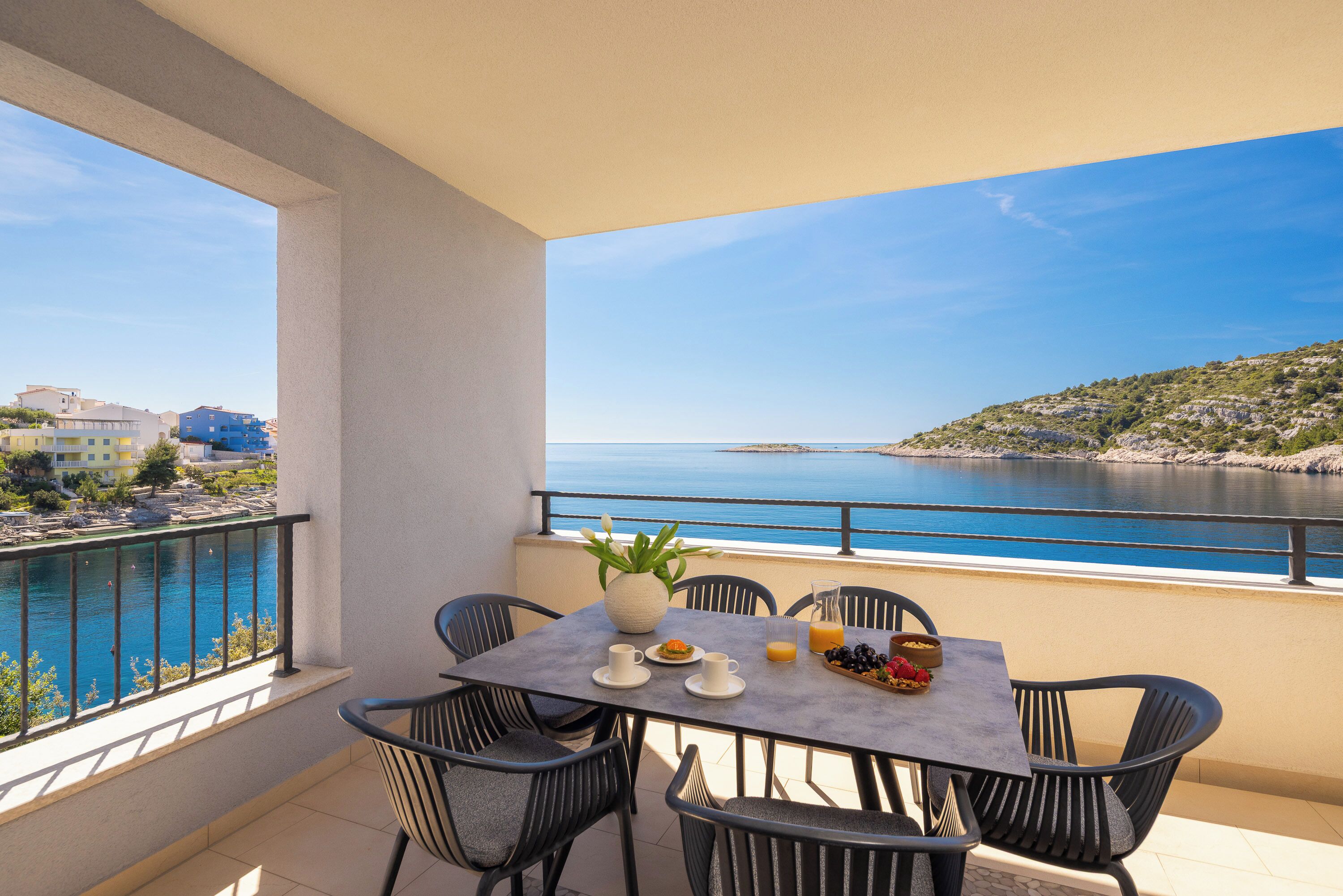 A balcony with a table set for breakfast, chairs, a vase with flowers, and a view of the sea and buildings.