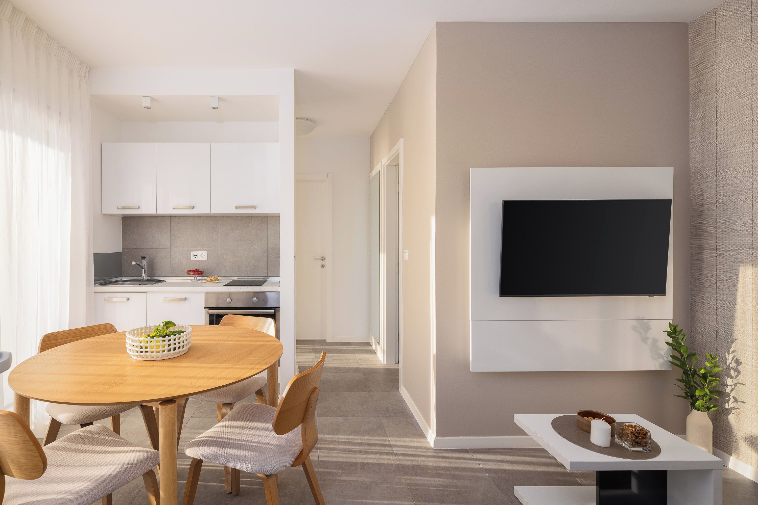 Une cuisine moderne avec des meubles blancs, un coin repas doté d’une table et de chaises en bois, et un téléviseur fixé au mur.