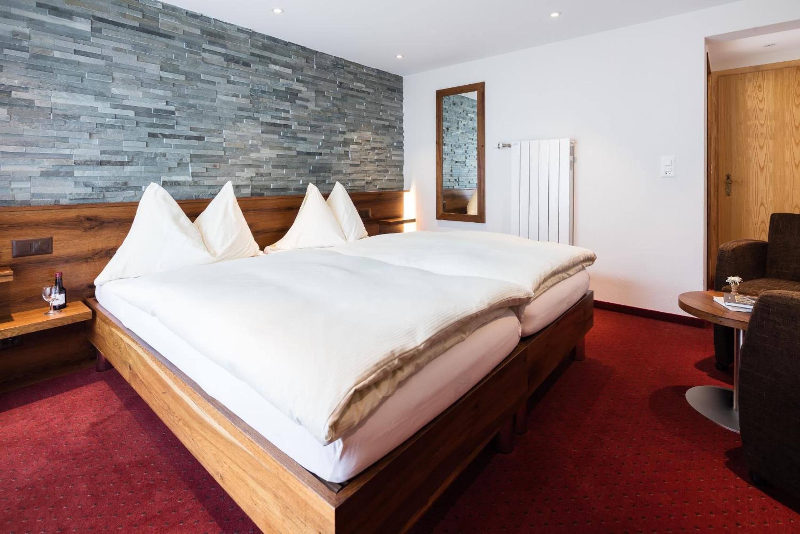 Habitación de hotel con una cama grande, cabecero de madera, pared de piedra, un espejo, un radiador y una pequeña mesa con una silla.