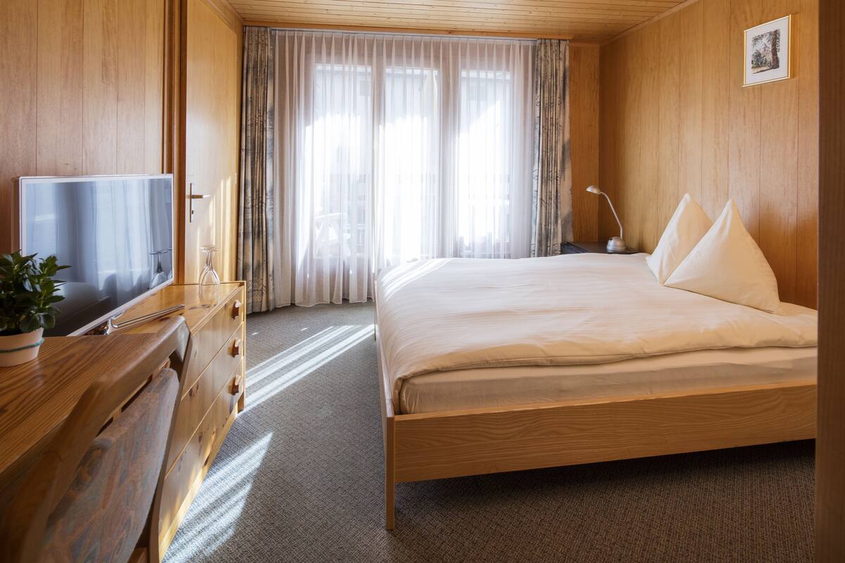 Een hotelkamer met een houten bed, een televisie, een bureau met lades, een lamp en een raam met gordijnen.
