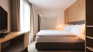 Habitación de hotel con cabecera de madera, cama con ropa blanca, escritorio con computadora y ventana con cortinas.