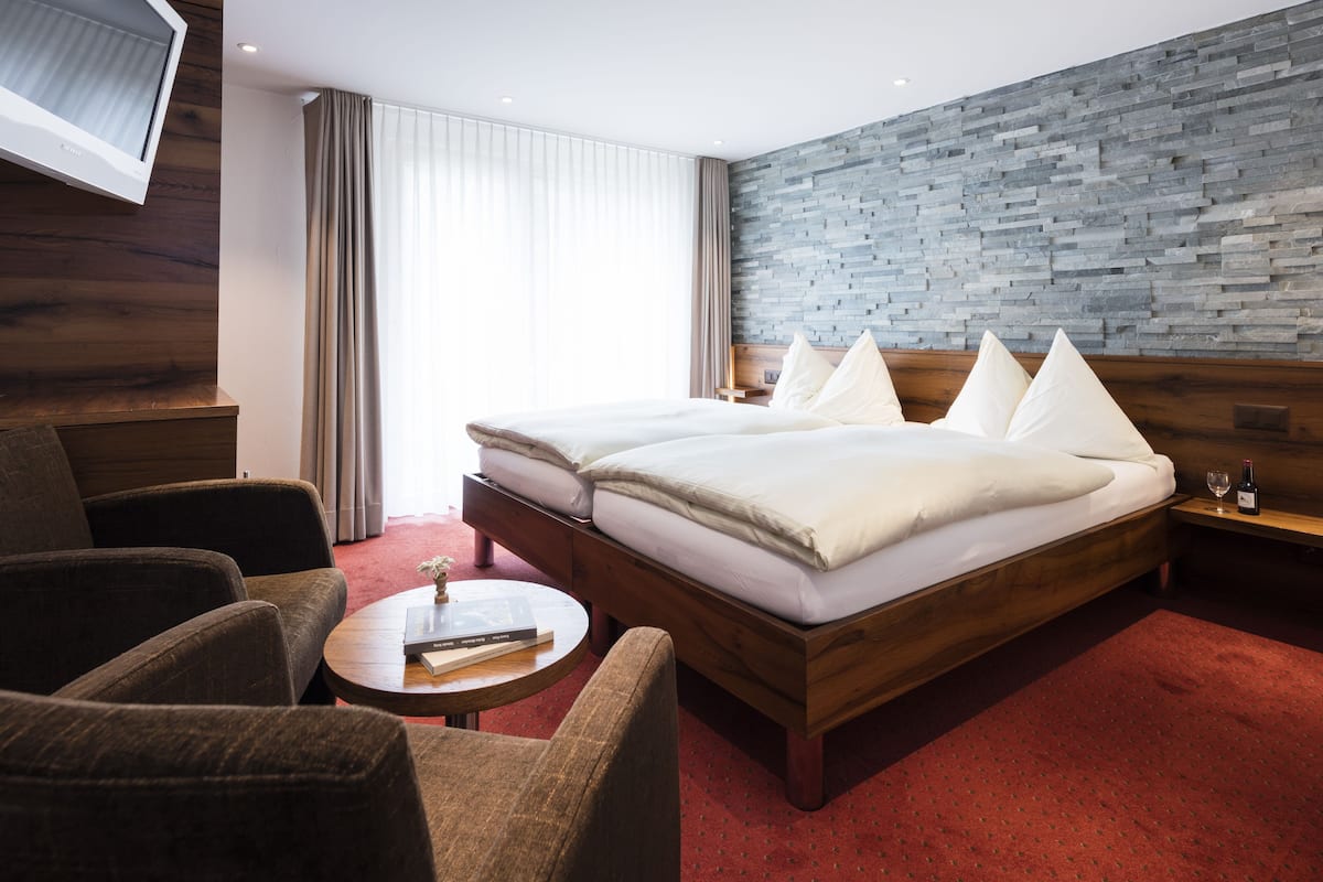 Een hotelkamer met een houten bed, twee fauteuils, een klein rond tafeltje en een aan de muur bevestigde televisie.