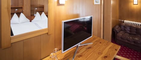 En TV med träram står på ett bord tillsammans med en bok och ett par glasögon. I rummet finns också en spegel och en lädersoffa. Väggarna är klädda med träpanel och det ligger en röd matta på golvet.