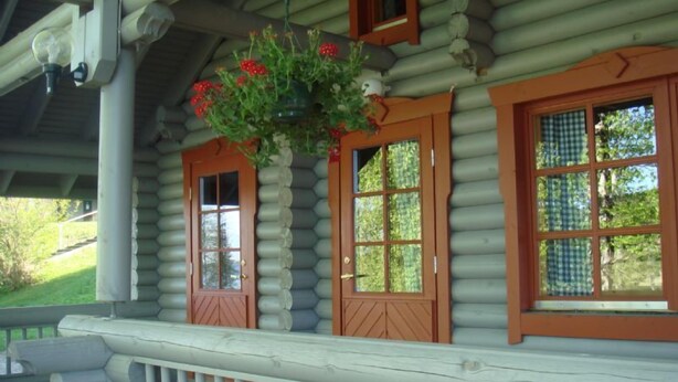 Une cabane en rondins avec une véranda, deux fenêtres dotées de volets en bois et un panier à fleurs suspendu.