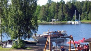 Un parc au bord du lac, avec une aire de jeux comprenant des balançoires, un bateau amarré sur l’eau et un espace pique-nique équipé de parasols.