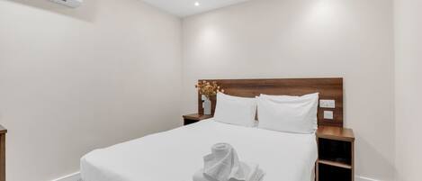 A neatly made bed with a white comforter and pillows, a wooden headboard, a nightstand with a vase of flowers, and a rolled towel on the bed.