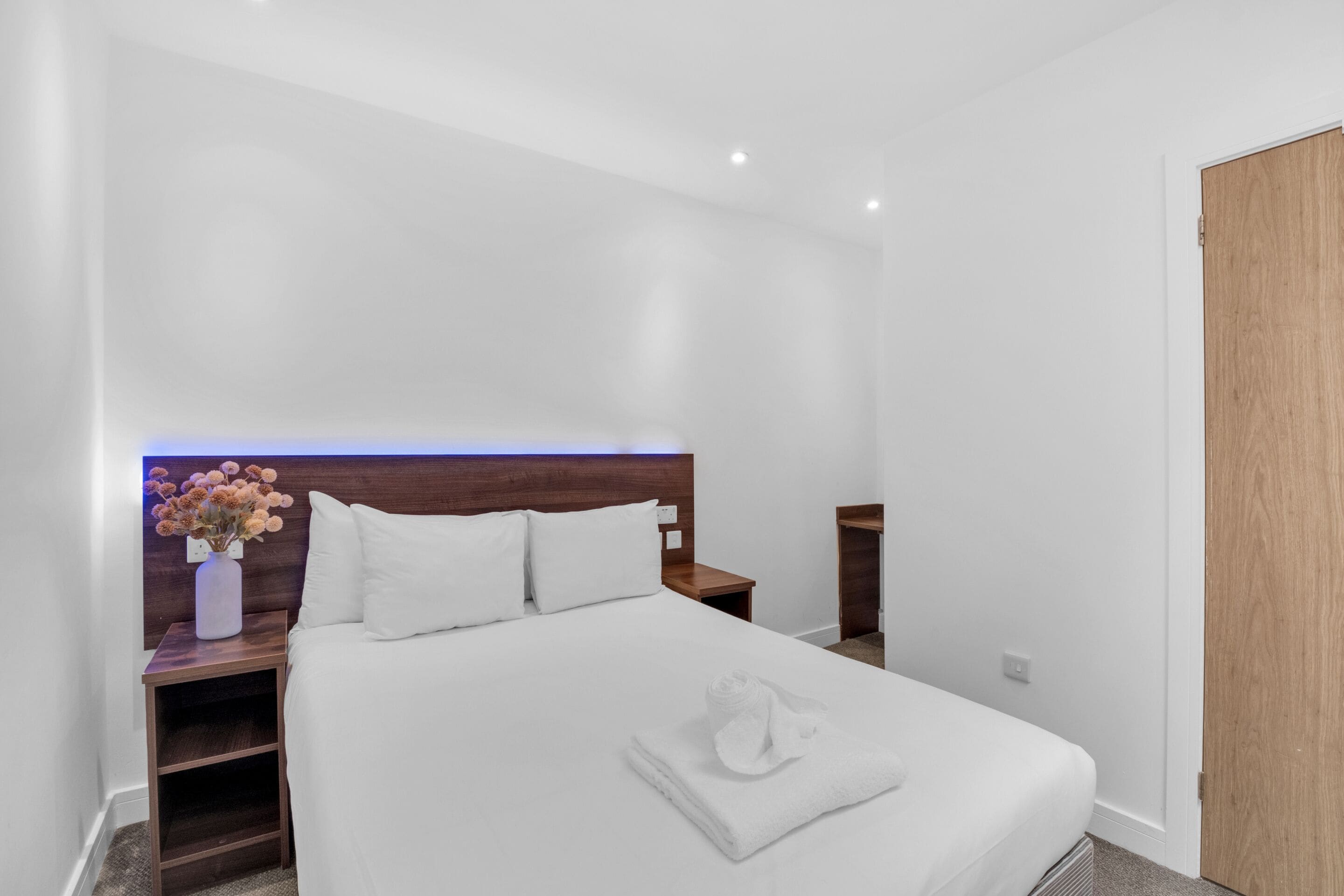 Une chambre d’hôtel moderne avec un lit soigneusement fait, une table de chevet en bois ornée d’un vase de fleurs et une porte en bois.