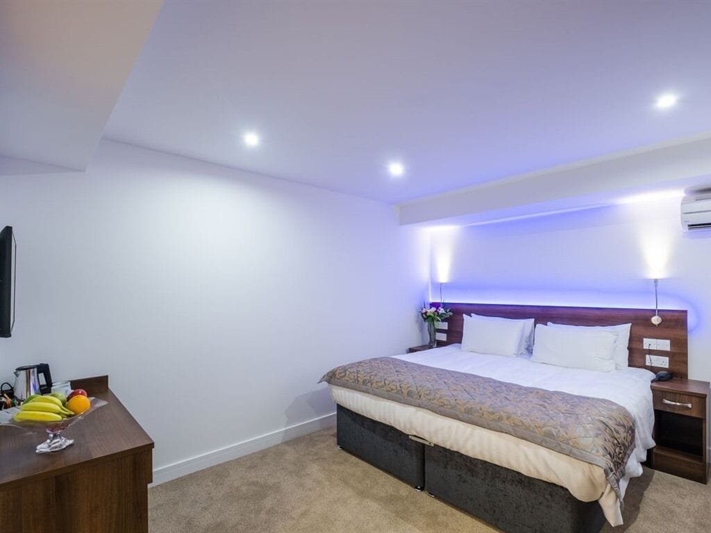 A hotel room with a bed, bedside tables, a desk with fruit, and a wall-mounted air conditioner.