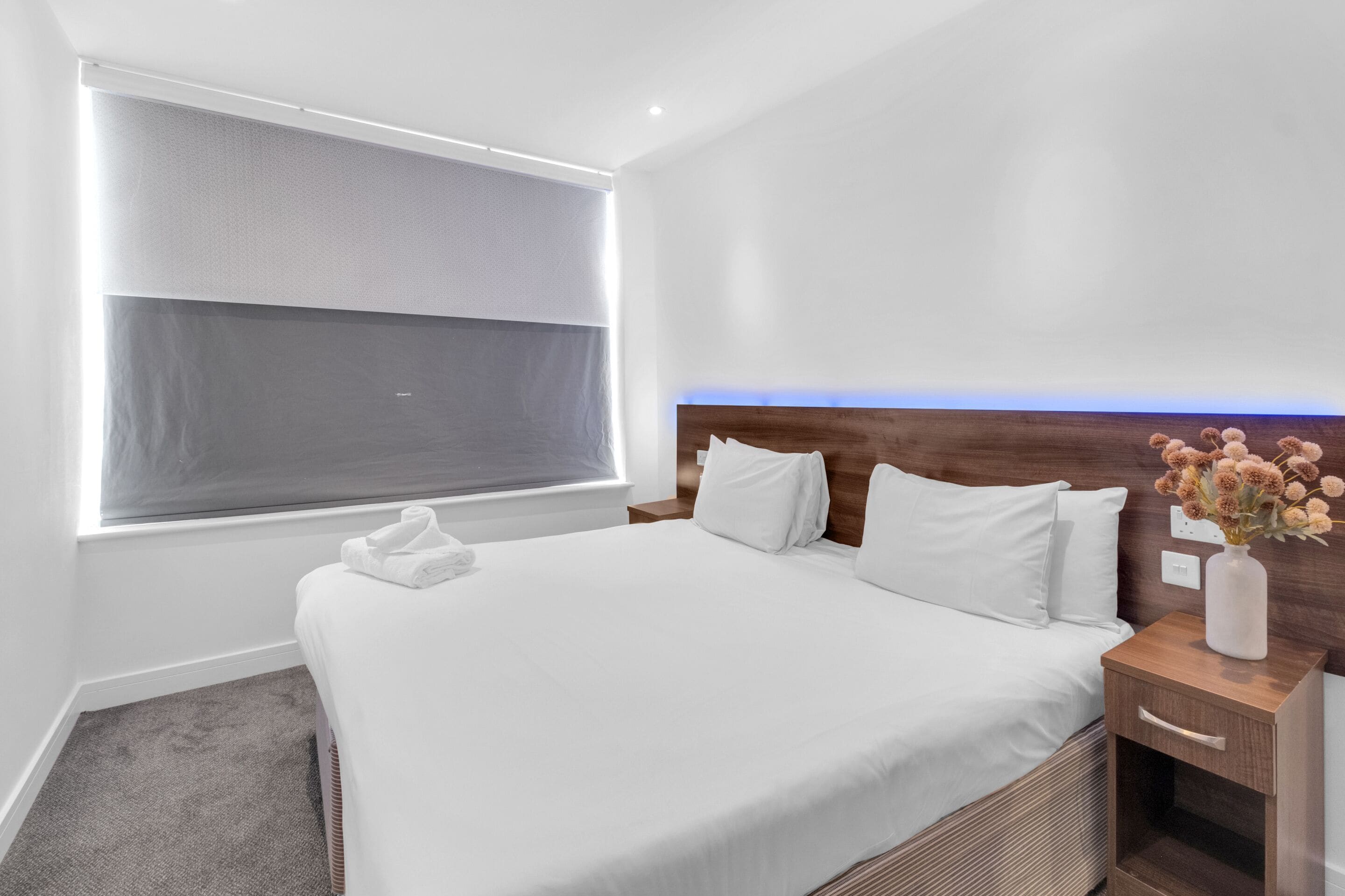 A modern hotel room with a large bed, a bedside table, a nightstand, a window with blinds, and a vase with flowers.