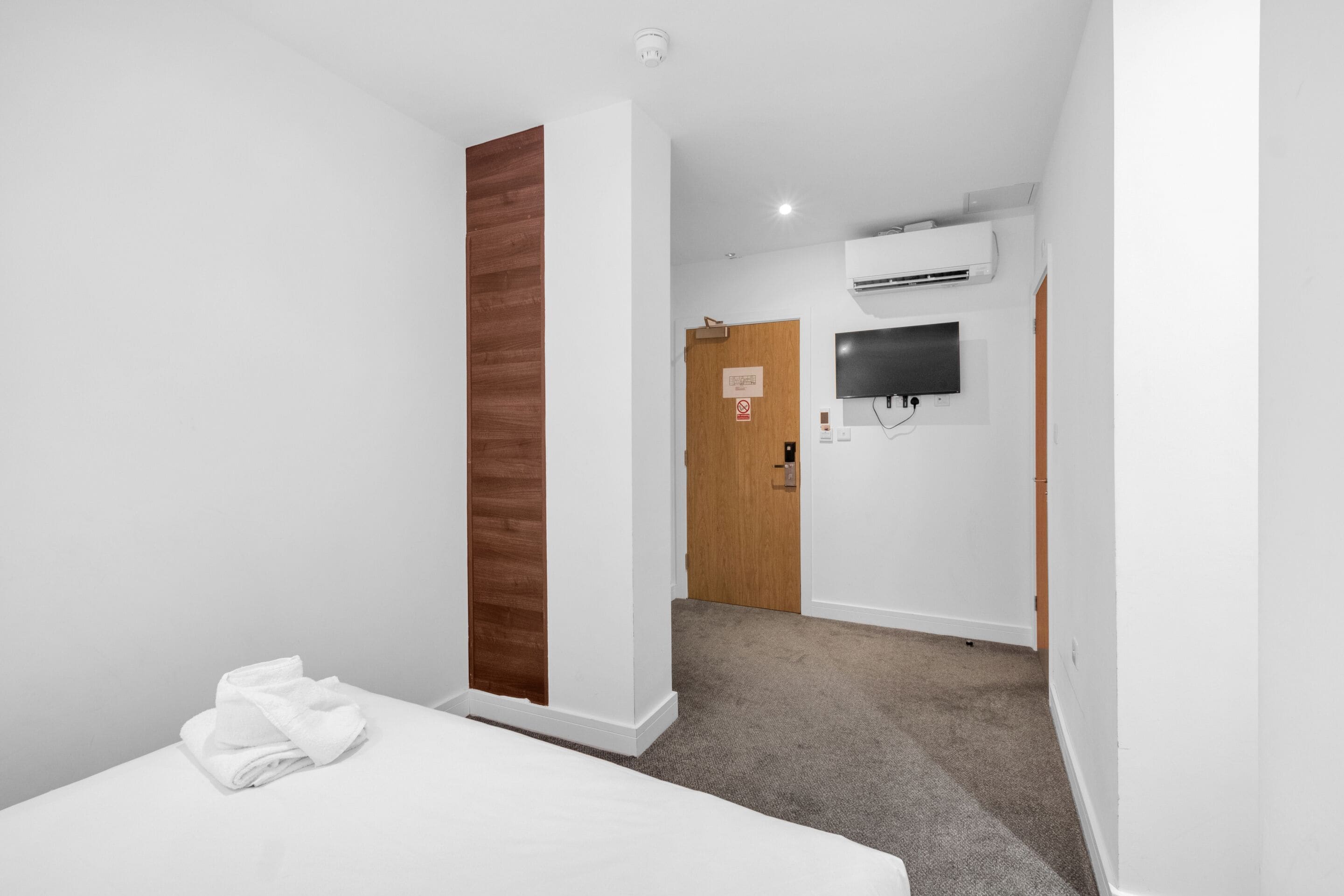 A hotel room with a bed, a wooden accent wall, a door, a mounted TV, and an air conditioning unit.