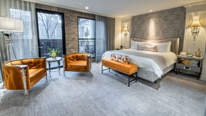 Quarto de hotel moderno com uma cama grande, área de estar com sofá e poltrona, uma mesinha e vista para o exterior por grandes janelas.