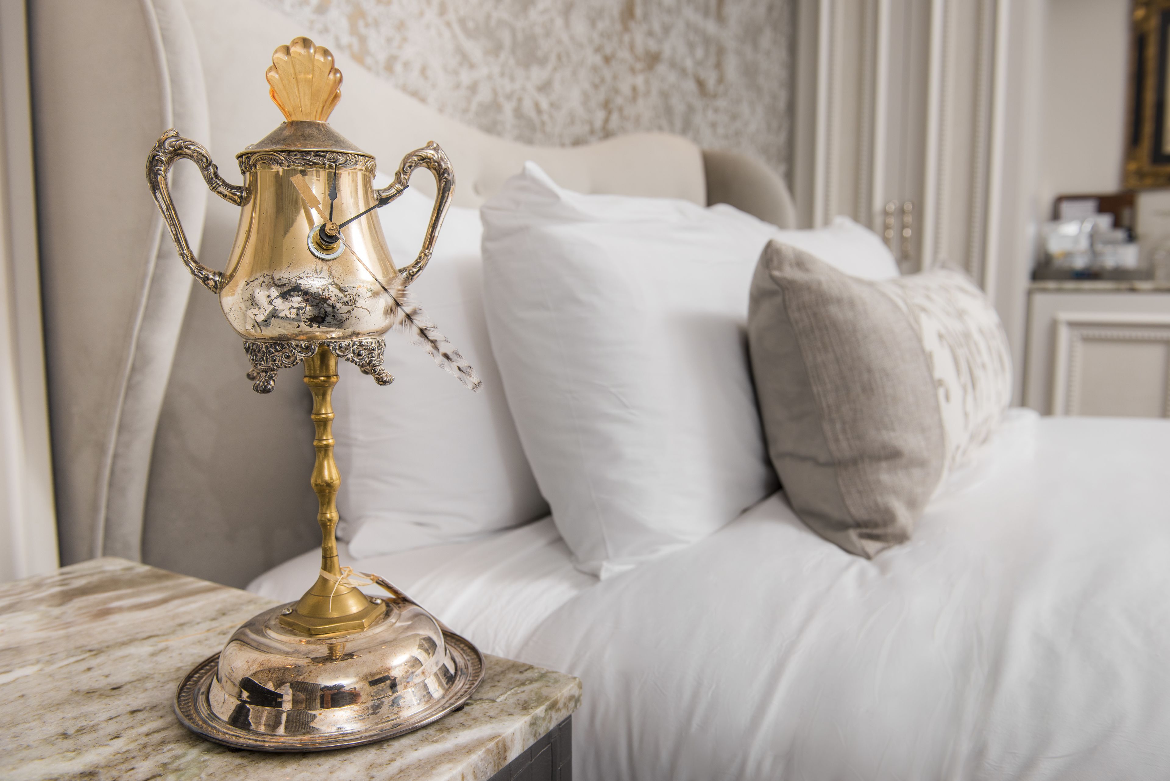 Uma cama com roupa de cama branca e um abajur decorativo prateado e dourado sobre uma superfície de mármore.