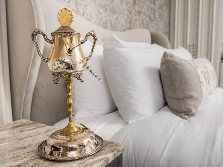 Uma cama com roupa de cama branca e um abajur decorativo prateado e dourado sobre uma superfície de mármore.