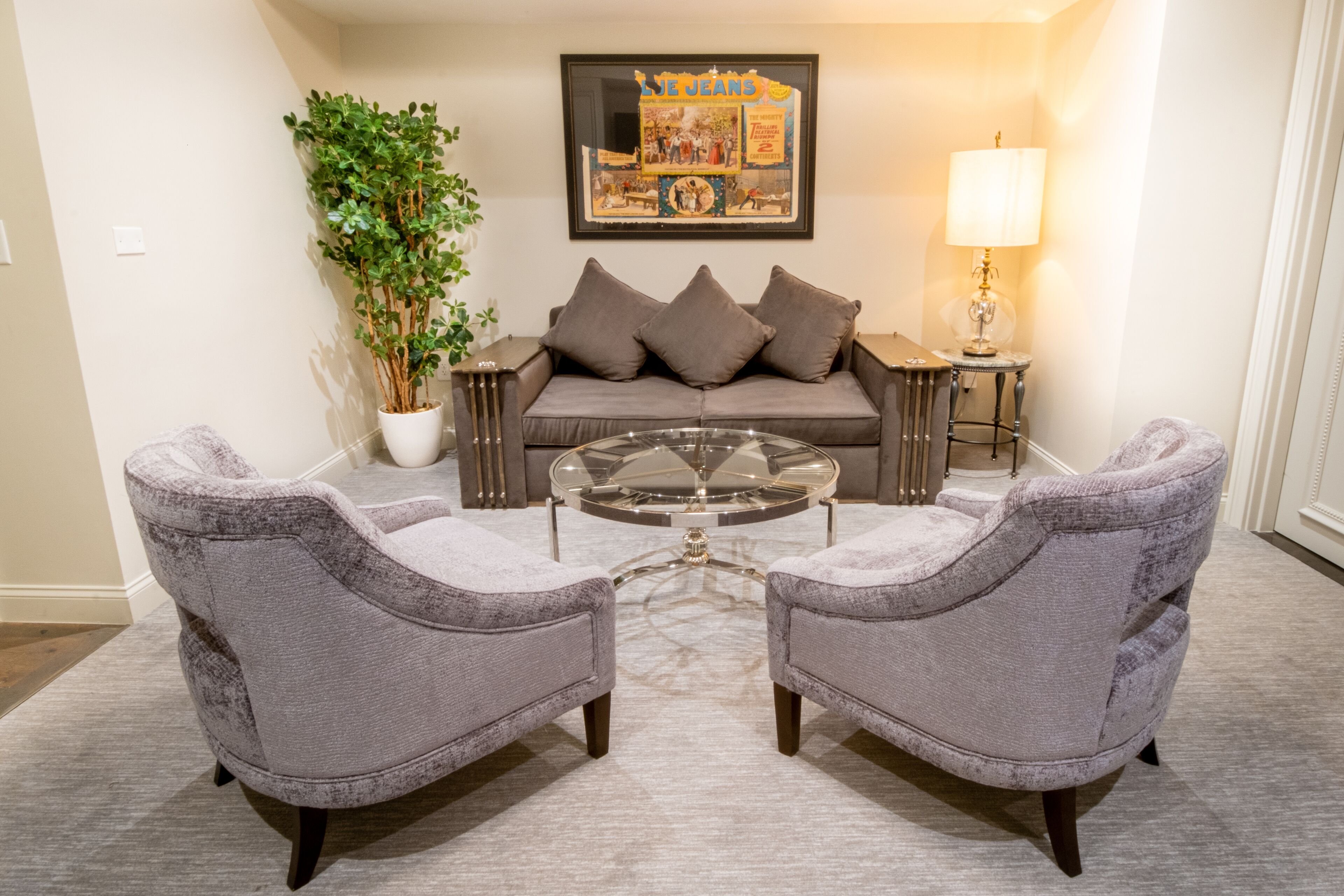 A living room with a grey sofa, two armchairs, a glass coffee table, a potted plant, a framed picture, and a lamp.